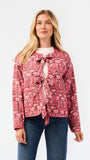 A woman modeling a pink floral patterned jacket with a tie-front detail and front pockets, paired with a white top and blue jeans, against a plain white studio background.