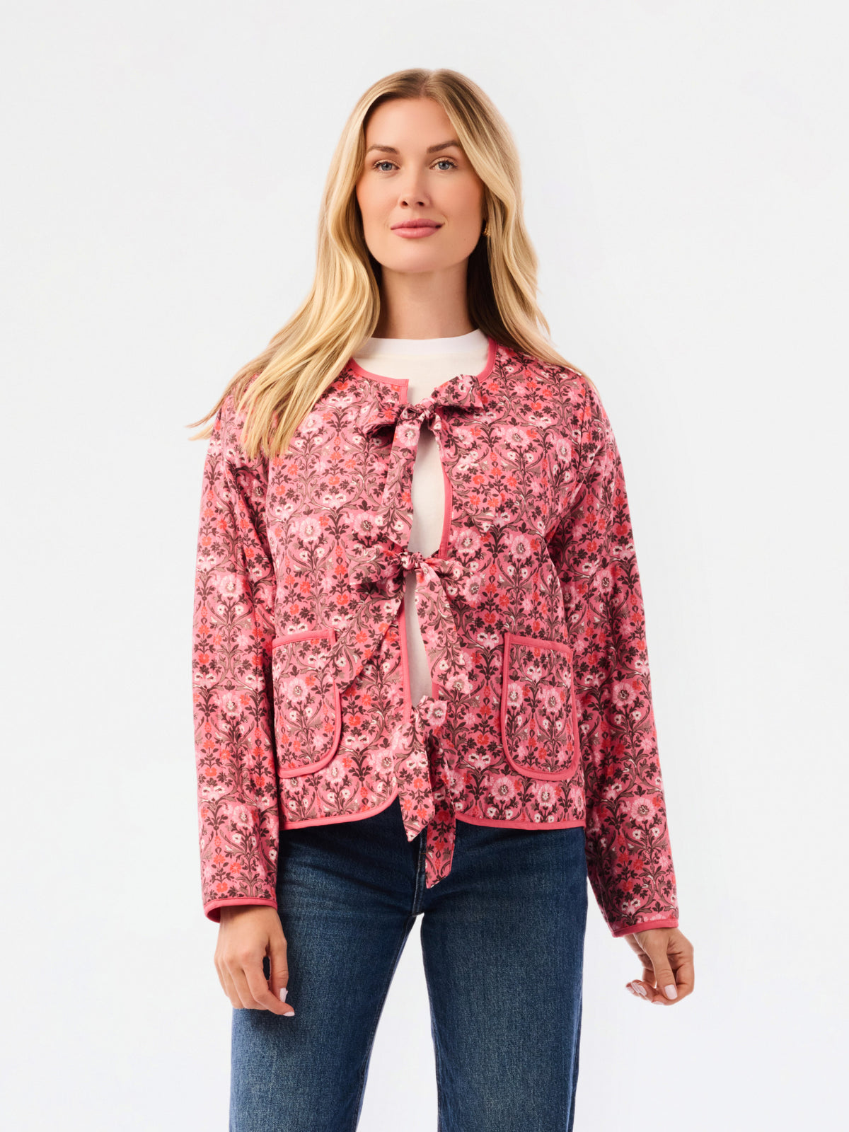 A woman modeling a pink floral patterned jacket with a tie-front detail and front pockets, paired with a white top and blue jeans, against a plain white studio background.