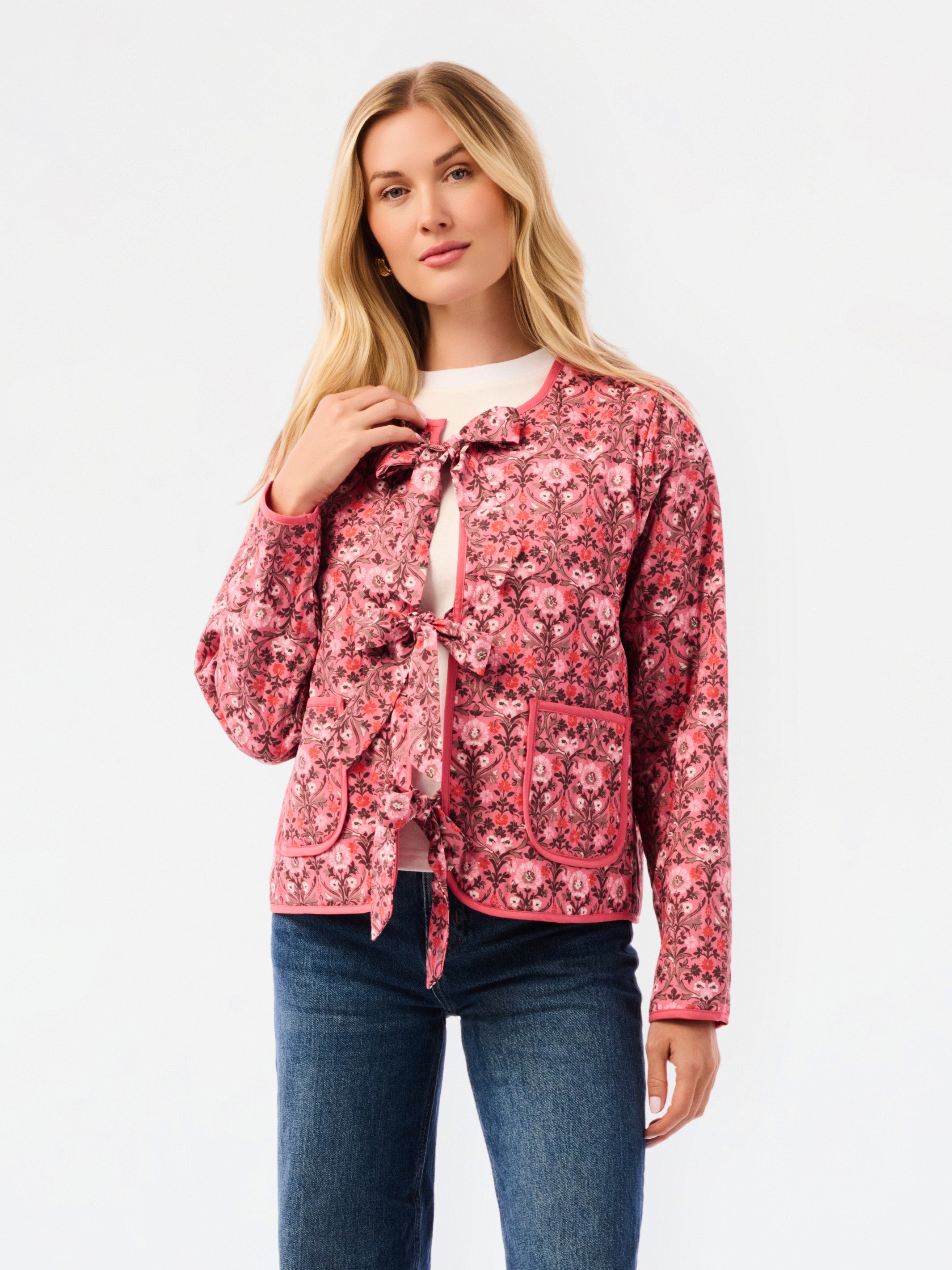A woman modeling a pink floral patterned jacket with a tie-front detail and front pockets, paired with a white top and blue jeans, against a plain white studio background.