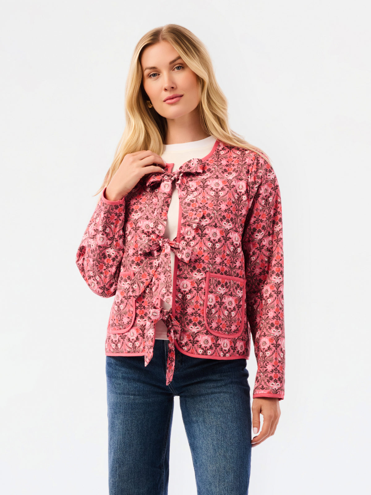 A woman modeling a pink floral patterned jacket with a tie-front detail and front pockets, paired with a white top and blue jeans, against a plain white studio background.