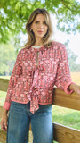 A woman stands outdoors leaning on a wooden fence, wearing a pink and red floral long-sleeve blouse with a tie-neck detail and blue jeans.
