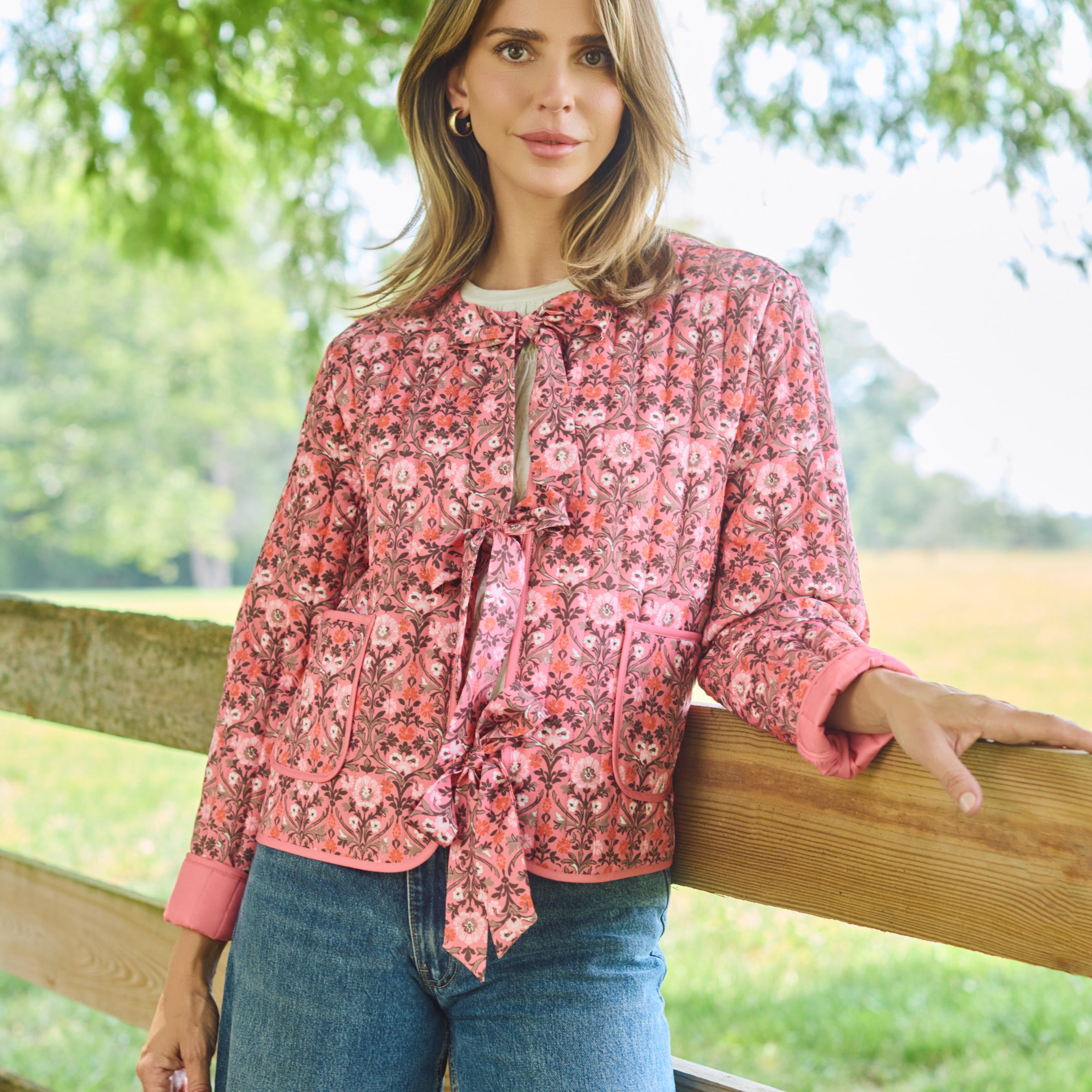 A woman stands outdoors leaning on a wooden fence, wearing a pink and red floral long-sleeve blouse with a tie-neck detail and blue jeans.