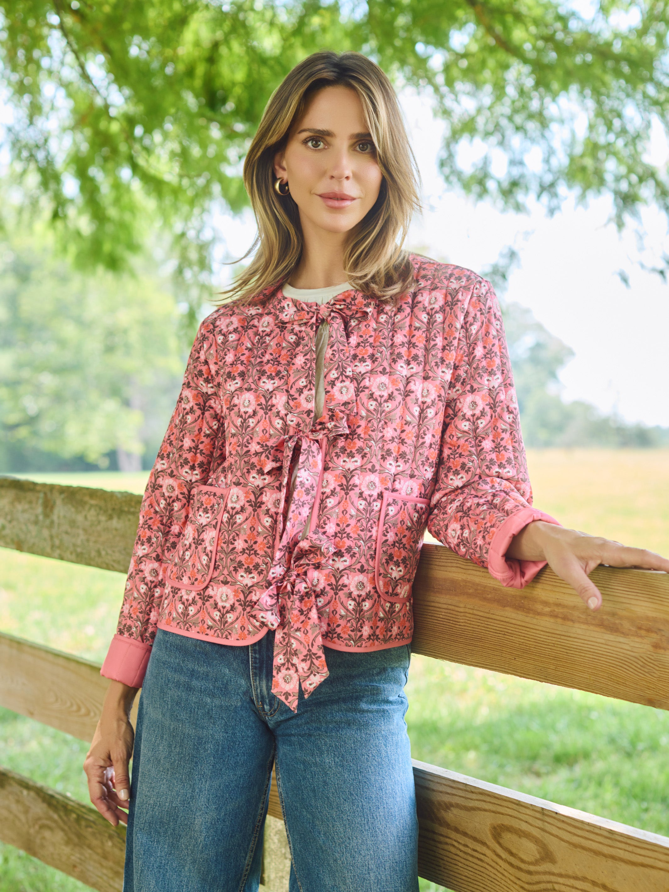 A woman stands outdoors leaning on a wooden fence, wearing a pink and red floral long-sleeve blouse with a tie-neck detail and blue jeans.
