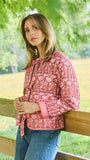 A woman wearing a pink floral long-sleeve jacket and blue jeans, leaning against a wooden fence in an outdoor park setting with green trees in the background.