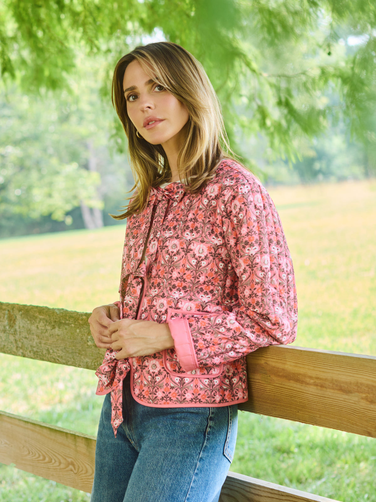 A woman wearing a pink floral long-sleeve jacket and blue jeans, leaning against a wooden fence in an outdoor park setting with green trees in the background.