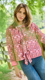 A woman wearing a pink floral print jacket with a tie waist and blue jeans, standing outdoors next to a wooden fence with green trees in the background.