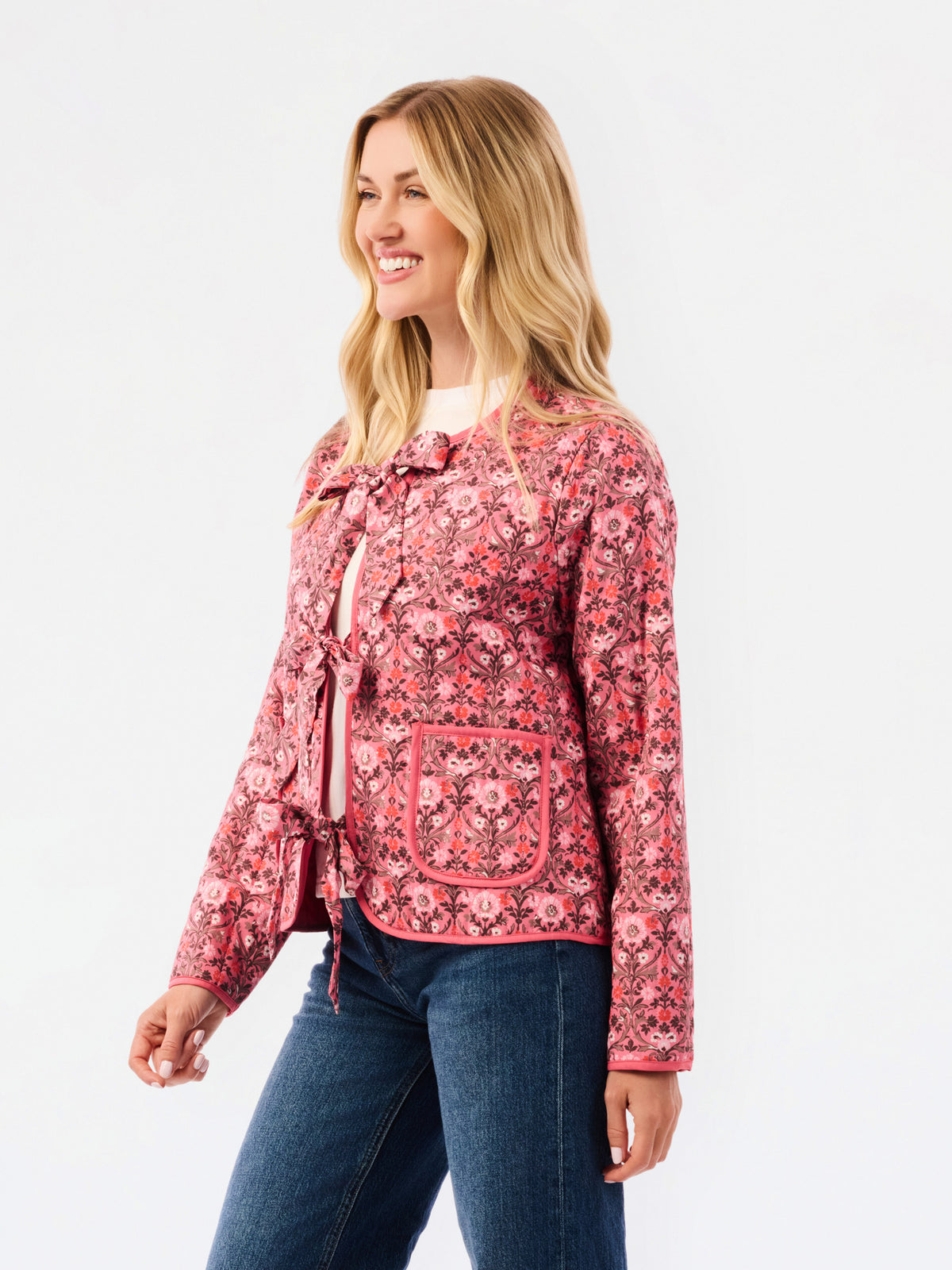 A woman modeling a pink and red floral patterned jacket with tie details and front pockets, paired with blue jeans, against a plain white studio background.