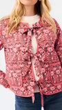 A woman modeling a pink floral patterned jacket with a tie-front detail and front pockets, paired with a white top and blue jeans, against a plain white studio background.
