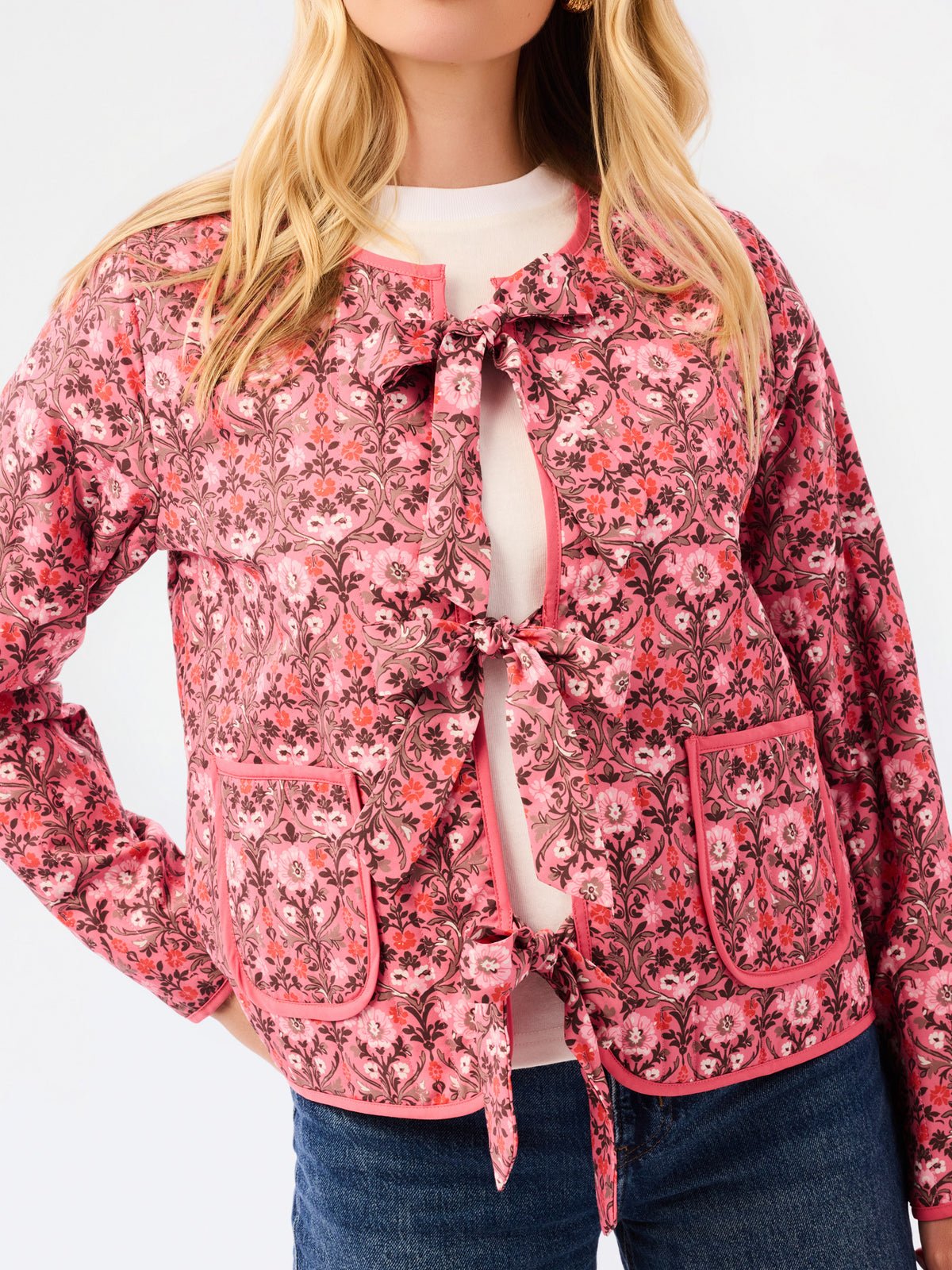 A woman modeling a pink floral patterned jacket with a tie-front detail and front pockets, paired with a white top and blue jeans, against a plain white studio background.