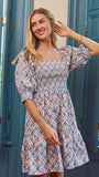 A woman wearing a short puff-sleeve dress with a blue, pink, and beige geometric pattern, standing in front of teal shutters.