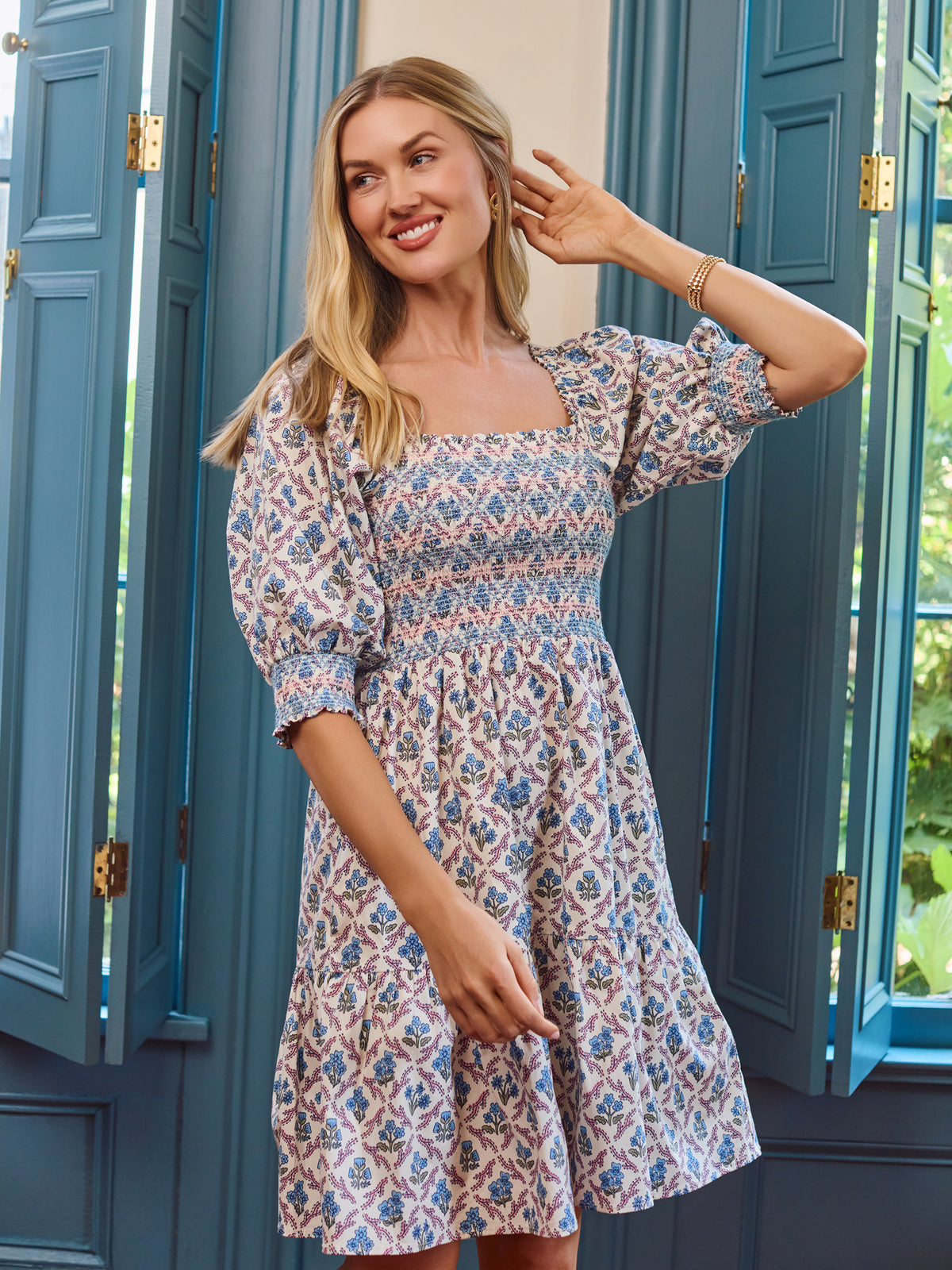 A woman wearing a short puff-sleeve dress with a blue, pink, and beige geometric pattern, standing in front of teal shutters.
