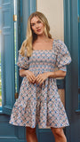 A woman wearing a short puff-sleeve dress with a blue, pink, and beige geometric pattern, standing in front of teal shutters.