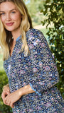 A woman wearing a blue and white patterned short-sleeve tunic dress, standing outdoors in front of a green hedge.
