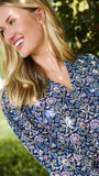 A woman wearing a blue and white patterned short-sleeve tunic dress, standing outdoors in front of a green hedge.