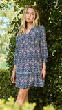 A woman wearing a blue and white patterned short-sleeve tunic dress, standing outdoors in front of a green hedge.