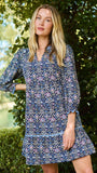 A woman wearing a blue and white patterned short-sleeve tunic dress, standing outdoors in front of a green hedge.
