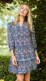 A woman wearing a blue and white patterned short-sleeve tunic dress, standing outdoors in front of a green hedge.