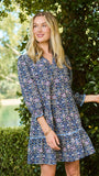 A woman wearing a blue and white patterned short-sleeve tunic dress, standing outdoors in front of a green hedge.