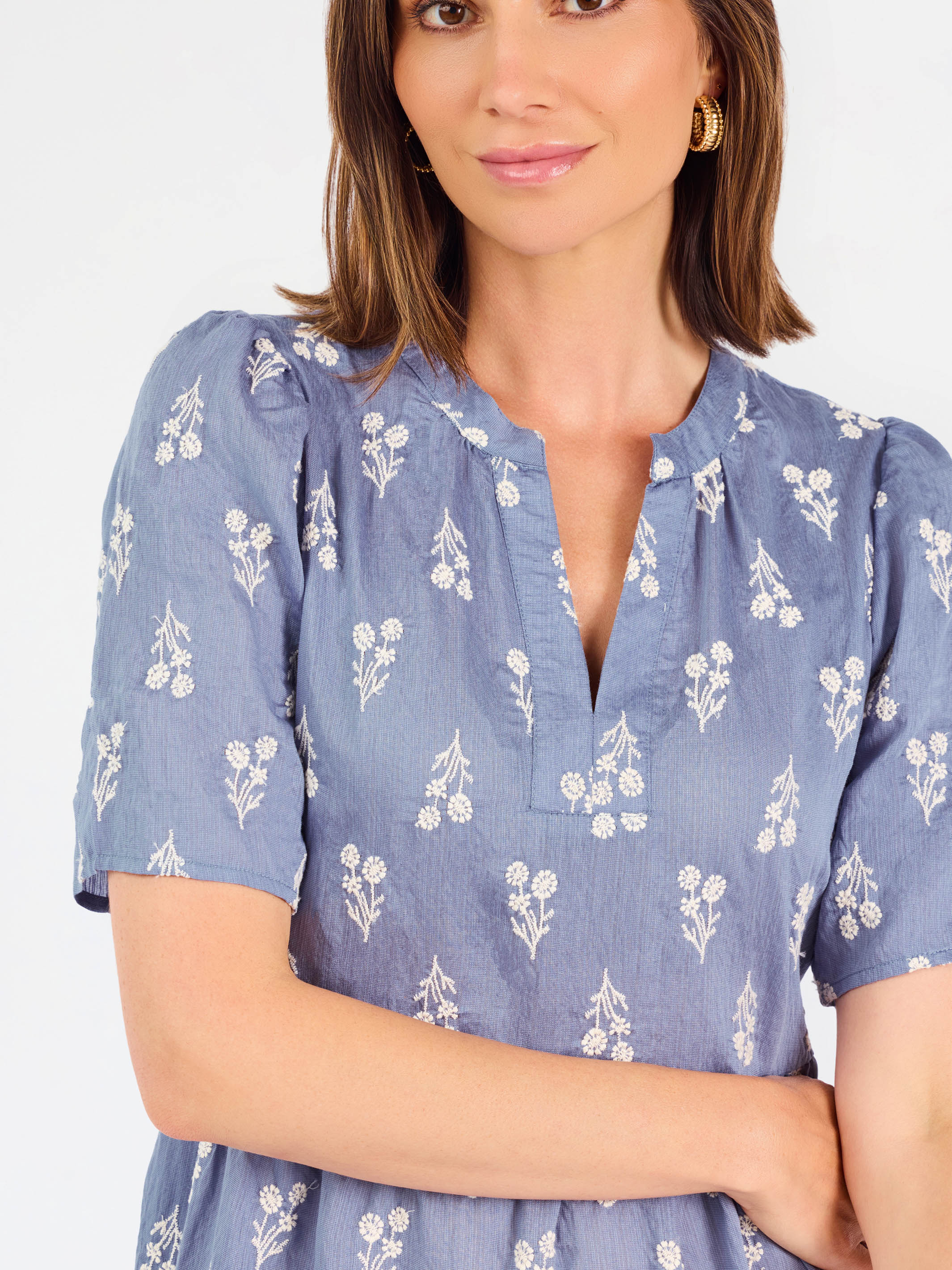 A woman wearing a dusty blue short-sleeve dress with a white floral print, featuring a split neckline and puff sleeves, shown against a plain white background.