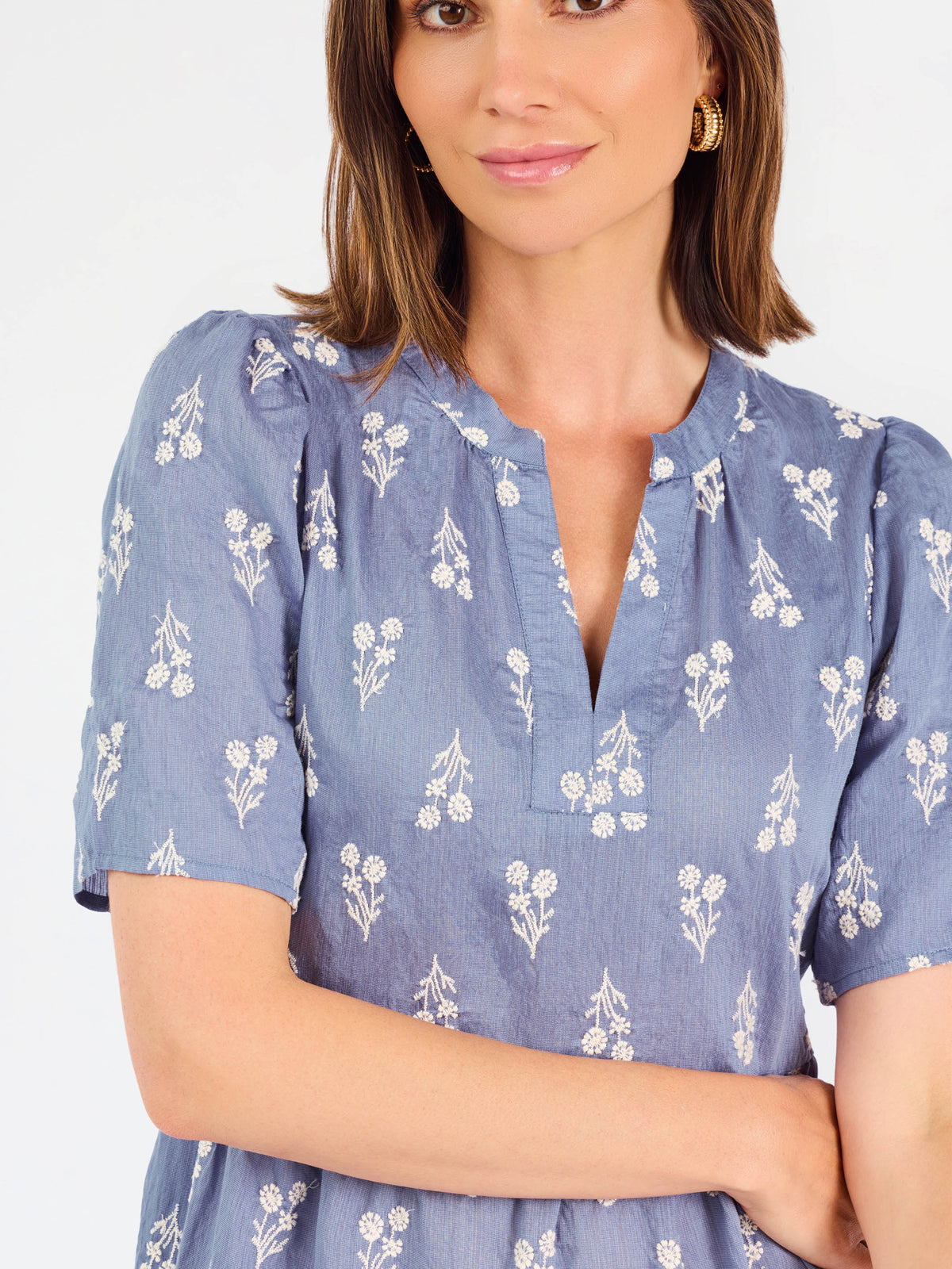 A woman wearing a dusty blue short-sleeve dress with a white floral print, featuring a split neckline and puff sleeves, shown against a plain white background.