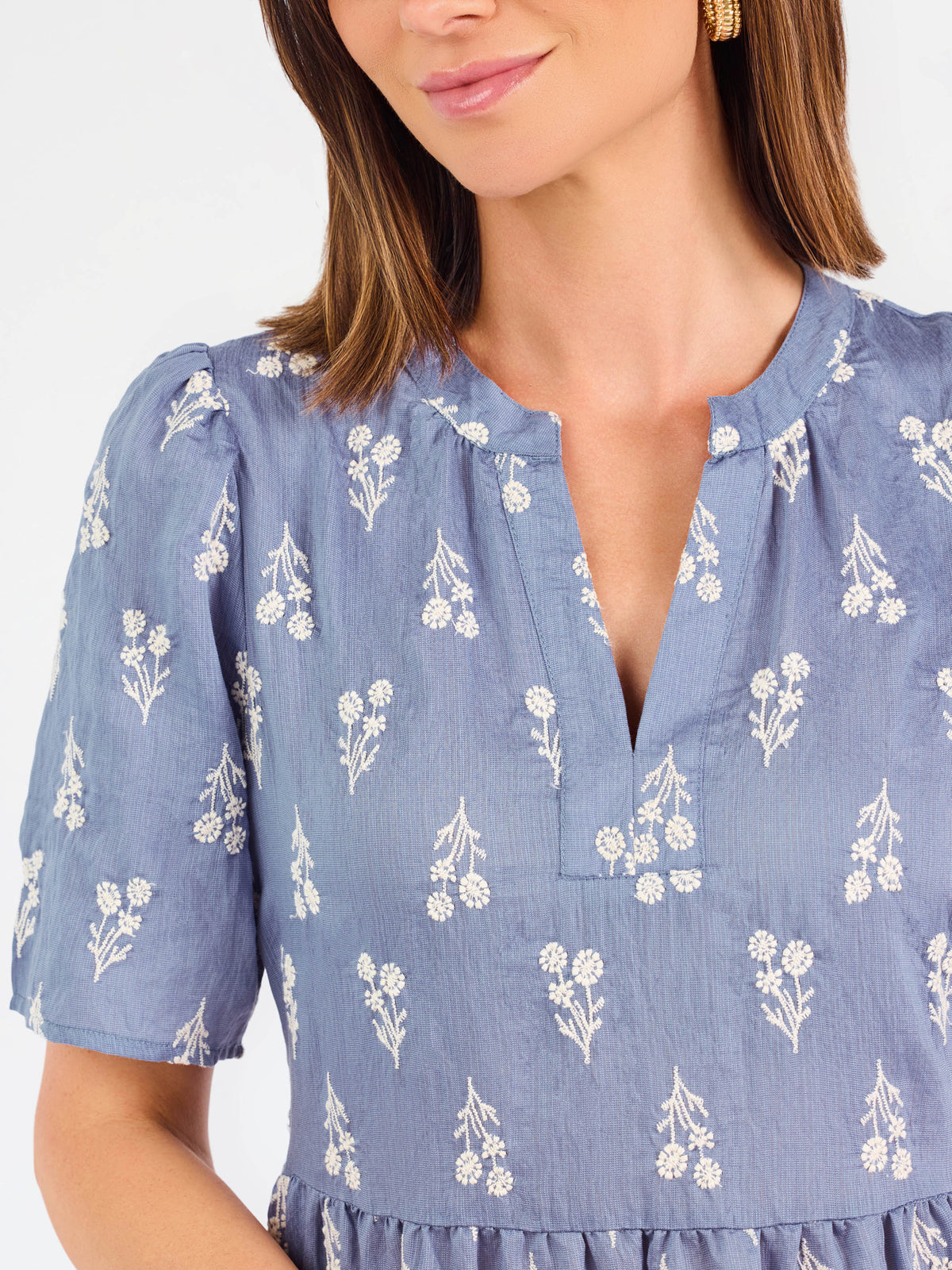 A woman wearing a dusty blue short-sleeve dress with a white floral print, featuring a split neckline and puff sleeves, shown against a plain white background.