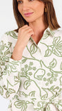 Woman wearing a white long-sleeve button-up dress with green floral embroidery, set against a plain light background.