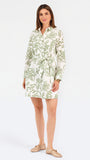 Woman wearing a white long-sleeve button-up dress with green floral embroidery, set against a plain light background.