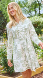 A woman wearing a long-sleeve white shirt dress with a green floral pattern, standing in a garden setting.