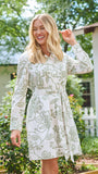 A woman wearing a long-sleeve white shirt dress with a green floral pattern, standing in a garden setting.