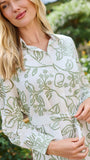 A woman wearing a long-sleeve white shirt dress with a green floral pattern, standing in a garden setting.