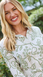A woman wearing a long-sleeve white shirt dress with a green floral pattern, standing in a garden setting.
