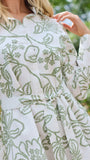 A woman wearing a long-sleeve white shirt dress with a green floral pattern, standing in a garden setting.