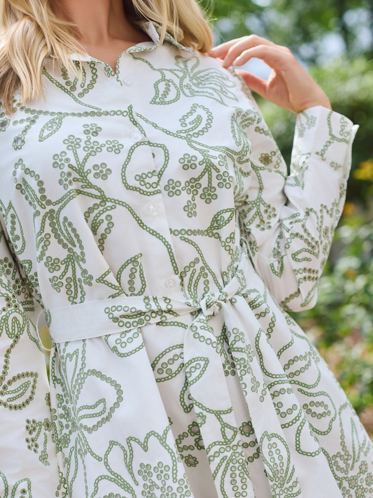 A woman wearing a long-sleeve white shirt dress with a green floral pattern, standing in a garden setting.