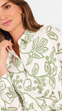 A close-up of a woman wearing a white long-sleeve button-up dress with green floral embroidery, set against a plain light background.