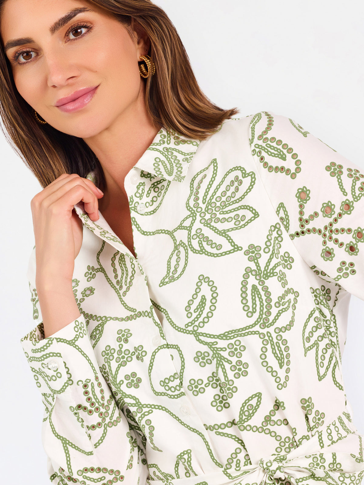 A close-up of a woman wearing a white long-sleeve button-up dress with green floral embroidery, set against a plain light background.