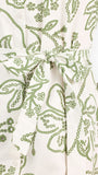Woman wearing a white long-sleeve button-up dress with green floral embroidery, set against a plain light background.
