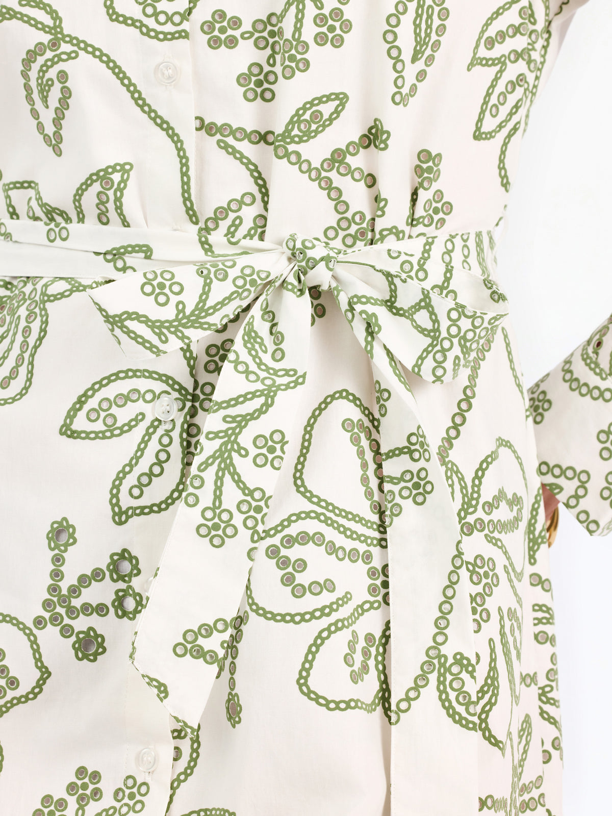 Woman wearing a white long-sleeve button-up dress with green floral embroidery, set against a plain light background.