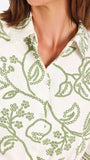 Woman wearing a white long-sleeve button-up dress with green floral embroidery, set against a plain light background.