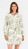 Woman wearing a white long-sleeve button-up dress with green floral embroidery, set against a plain light background.