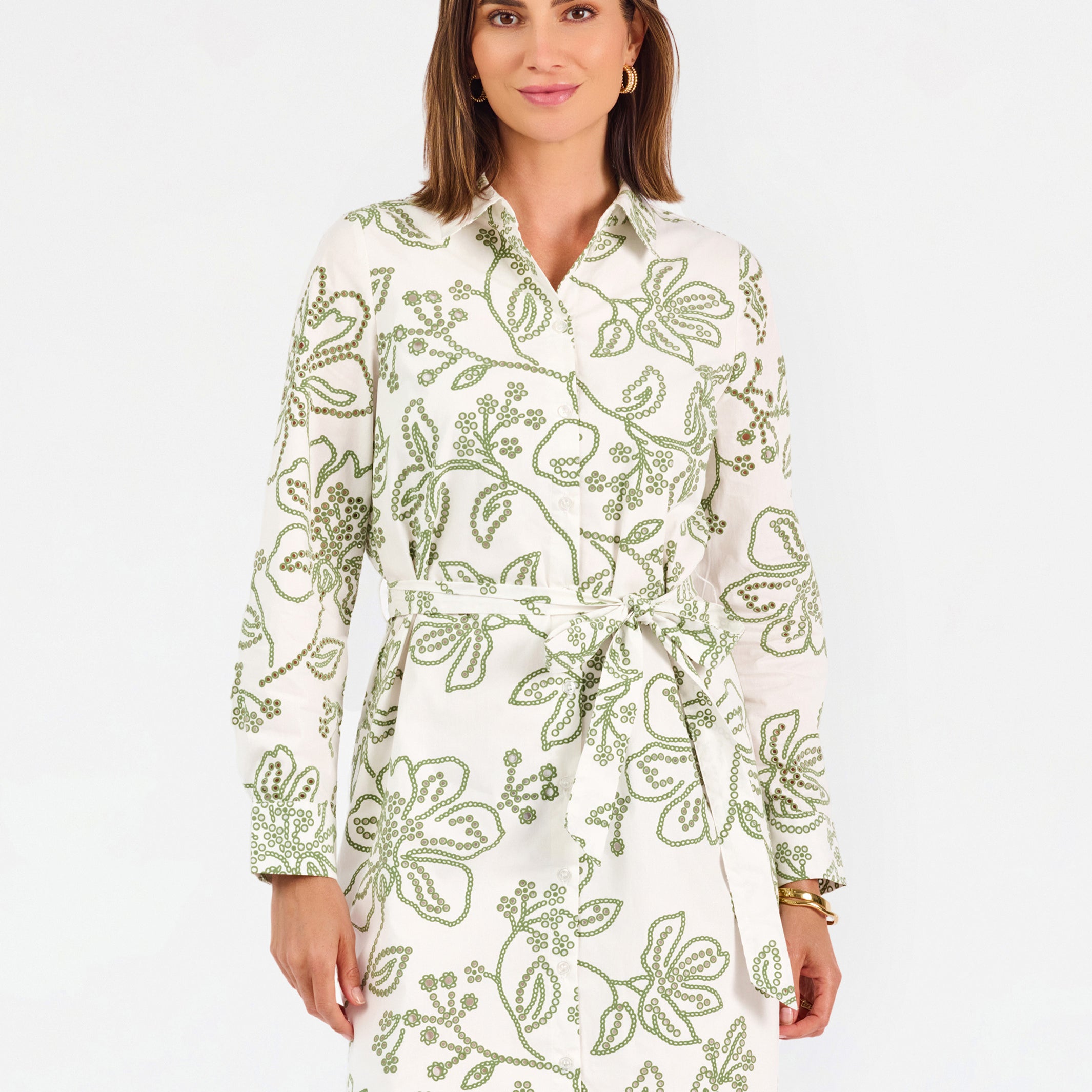 Woman wearing a white long-sleeve button-up dress with green floral embroidery, set against a plain light background.