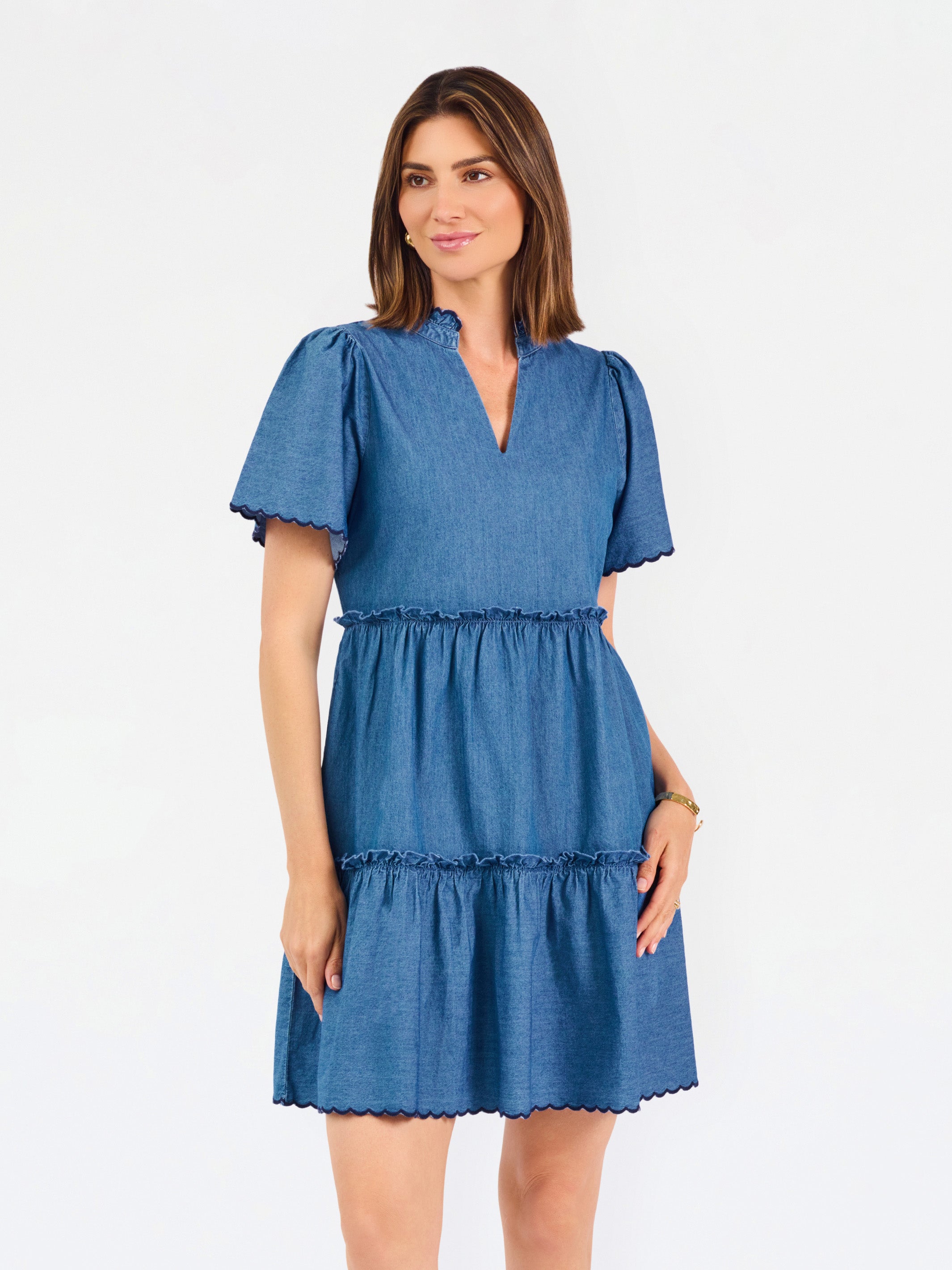 A woman modeling a short-sleeve tiered dress in medium blue chambray fabric against a plain white studio background.