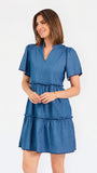 A woman modeling a short-sleeve tiered dress in medium blue chambray fabric against a plain white studio background.