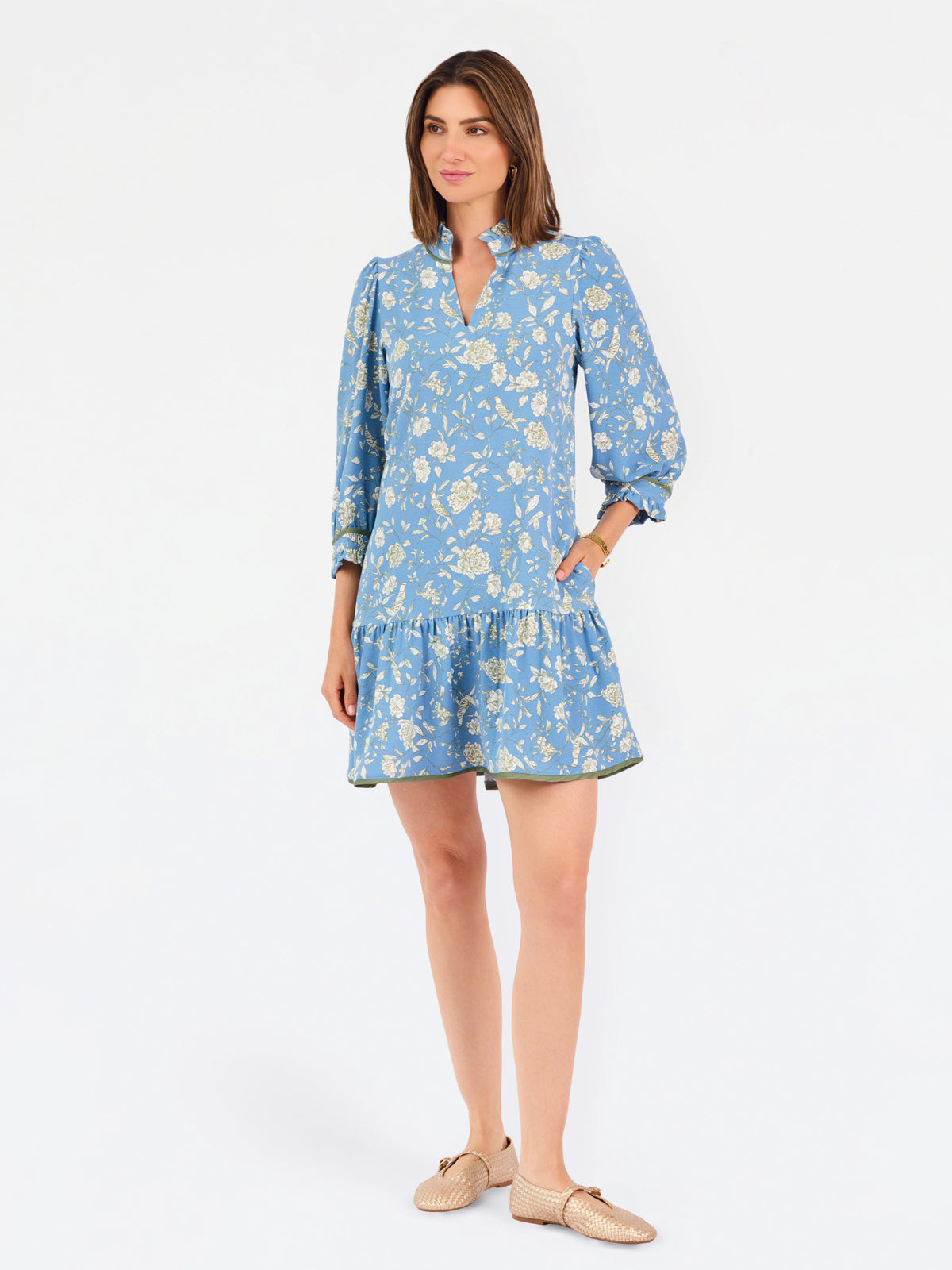A woman modeling a light blue long-sleeve mini dress with a cream floral print, standing against a plain white studio background.