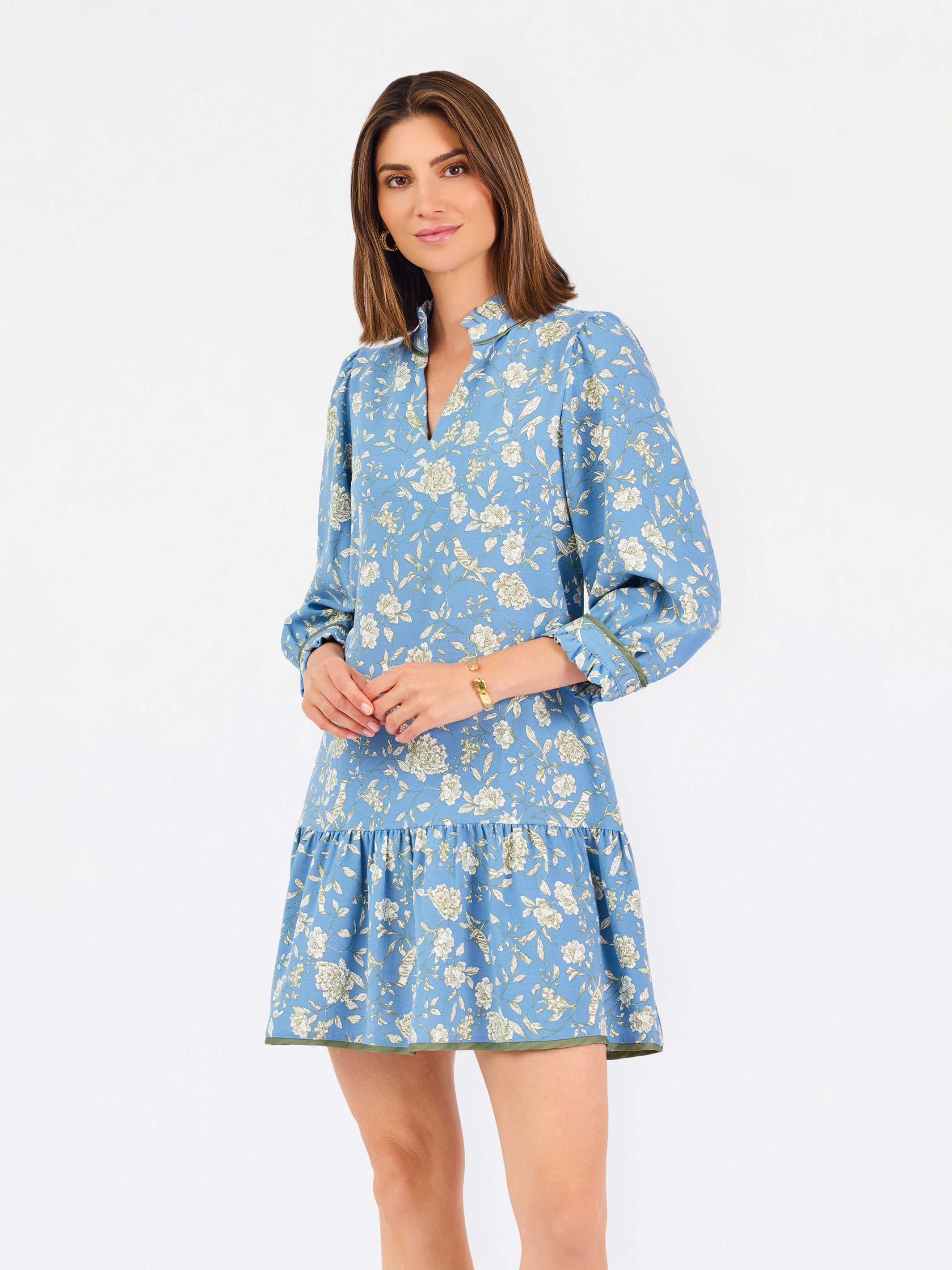 A woman modeling a light blue long-sleeve mini dress with a cream floral print, standing against a plain white studio background.