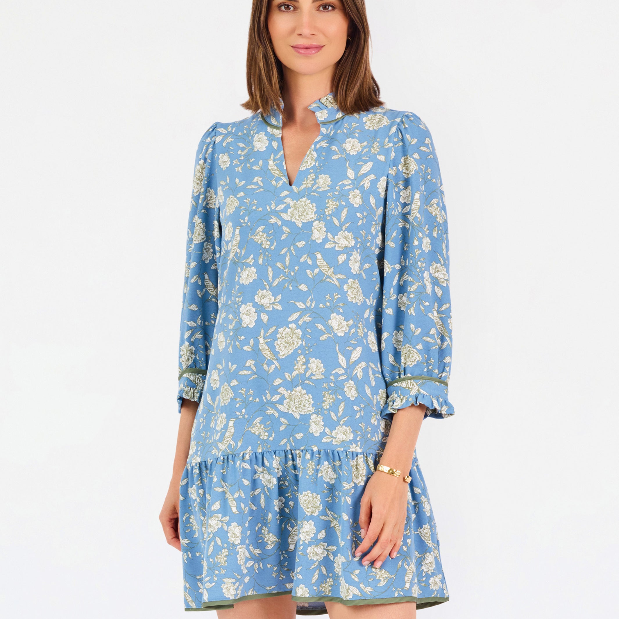 A woman modeling a light blue long-sleeve mini dress with a cream floral print, standing against a plain white studio background.