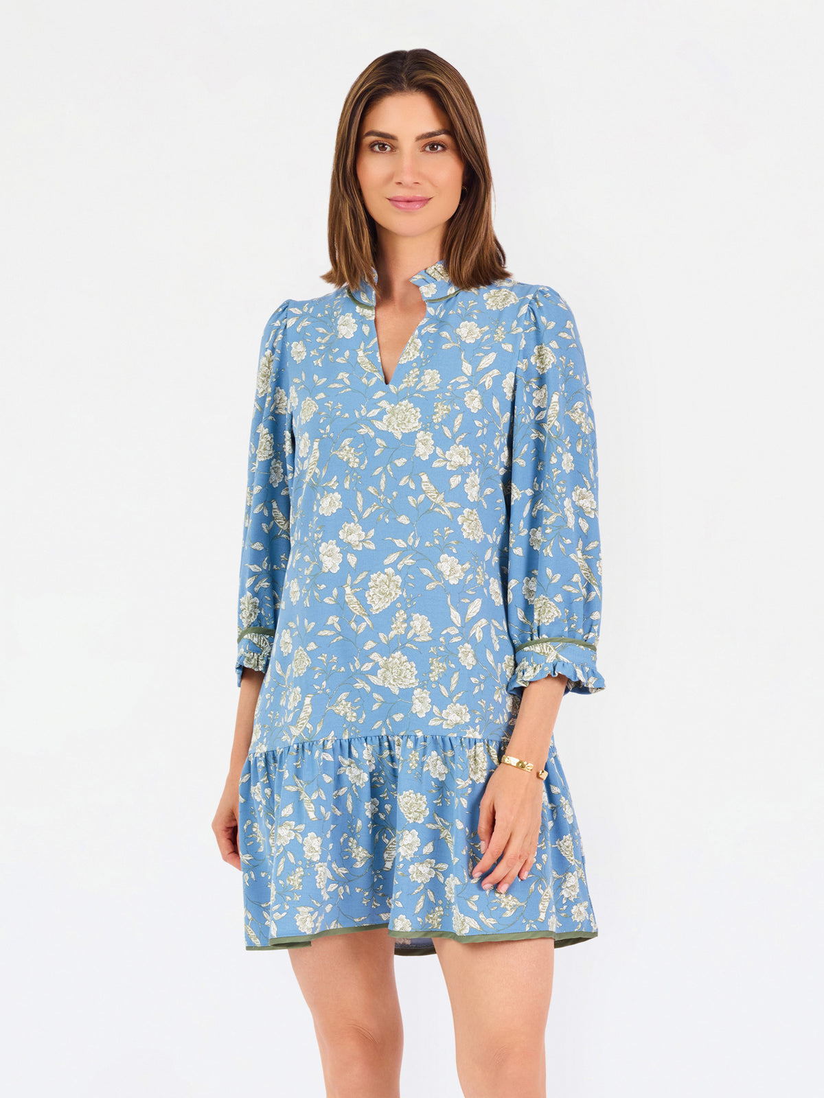 A woman modeling a light blue long-sleeve mini dress with a cream floral print, standing against a plain white studio background.