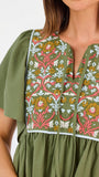 A woman modeling a short-sleeve olive green dress with a colorful floral embroidered yoke and a gathered waist, paired with beige flat shoes against a plain white background.