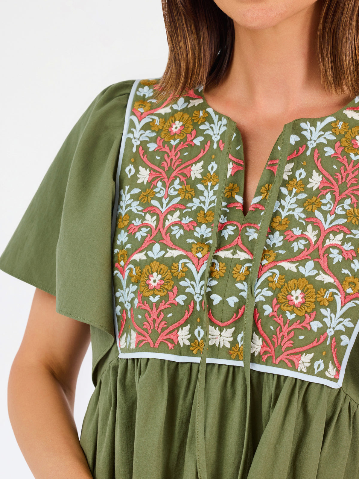 A woman modeling a short-sleeve olive green dress with a colorful floral embroidered yoke and a gathered waist, paired with beige flat shoes against a plain white background.