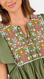 A woman modeling a short-sleeve olive green dress with a colorful floral embroidered yoke and a gathered waist, paired with beige flat shoes against a plain white background.
