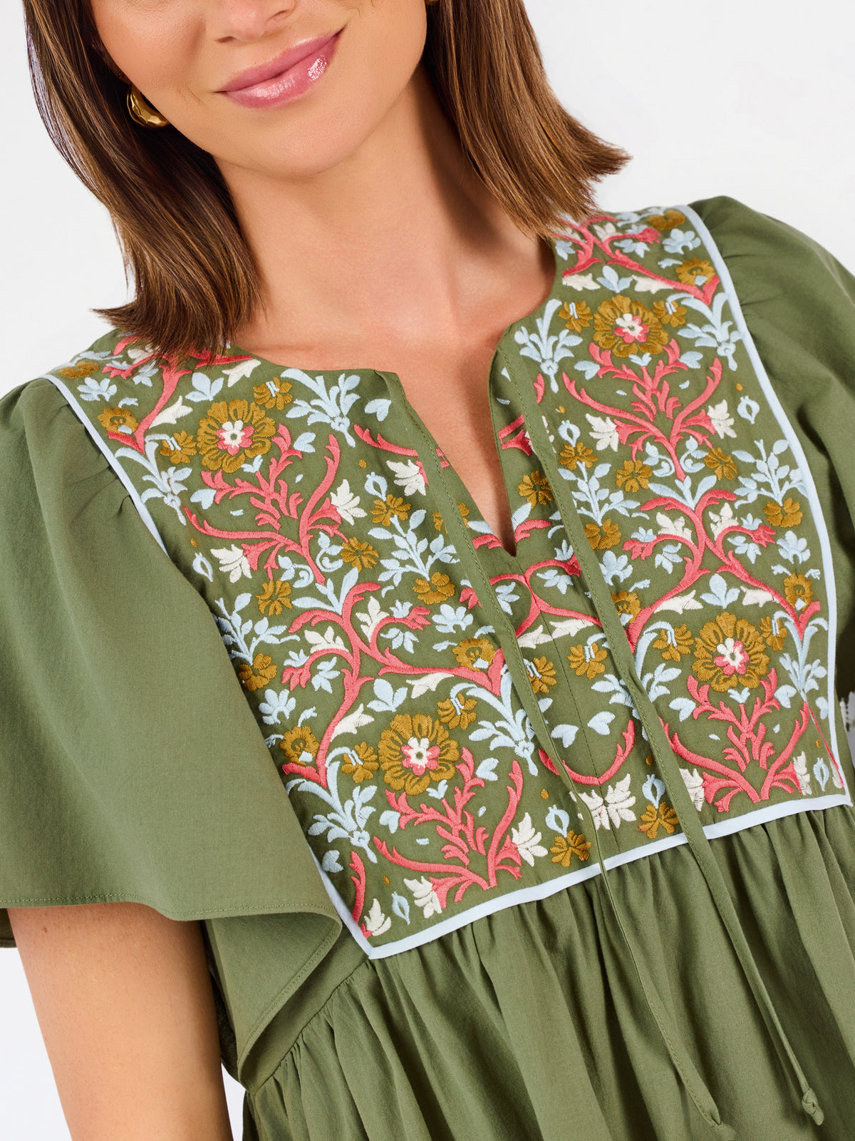 A woman modeling a short-sleeve olive green dress with a colorful floral embroidered yoke and a gathered waist, paired with beige flat shoes against a plain white background.